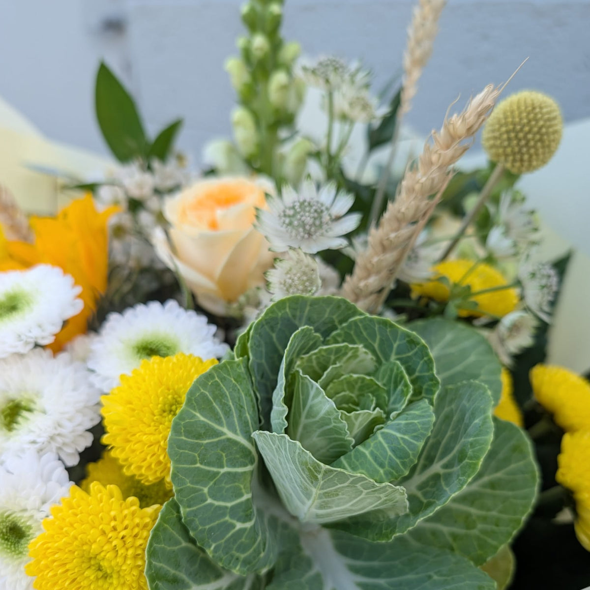 A close-up of a mixed floral arrangement featuring a large green ornamental cabbage flower, bright yellow chrysanthemums, white daisies, a pale peach rose, and dried wheat stems, wrapped in cream paper.