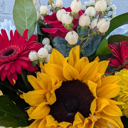 A close-up of a mixed flower arrangement featuring a large yellow sunflower in the foreground, red gerbera daisies on the sides, white bell-shaped flowers, and green foliage against a light blue textured wall.