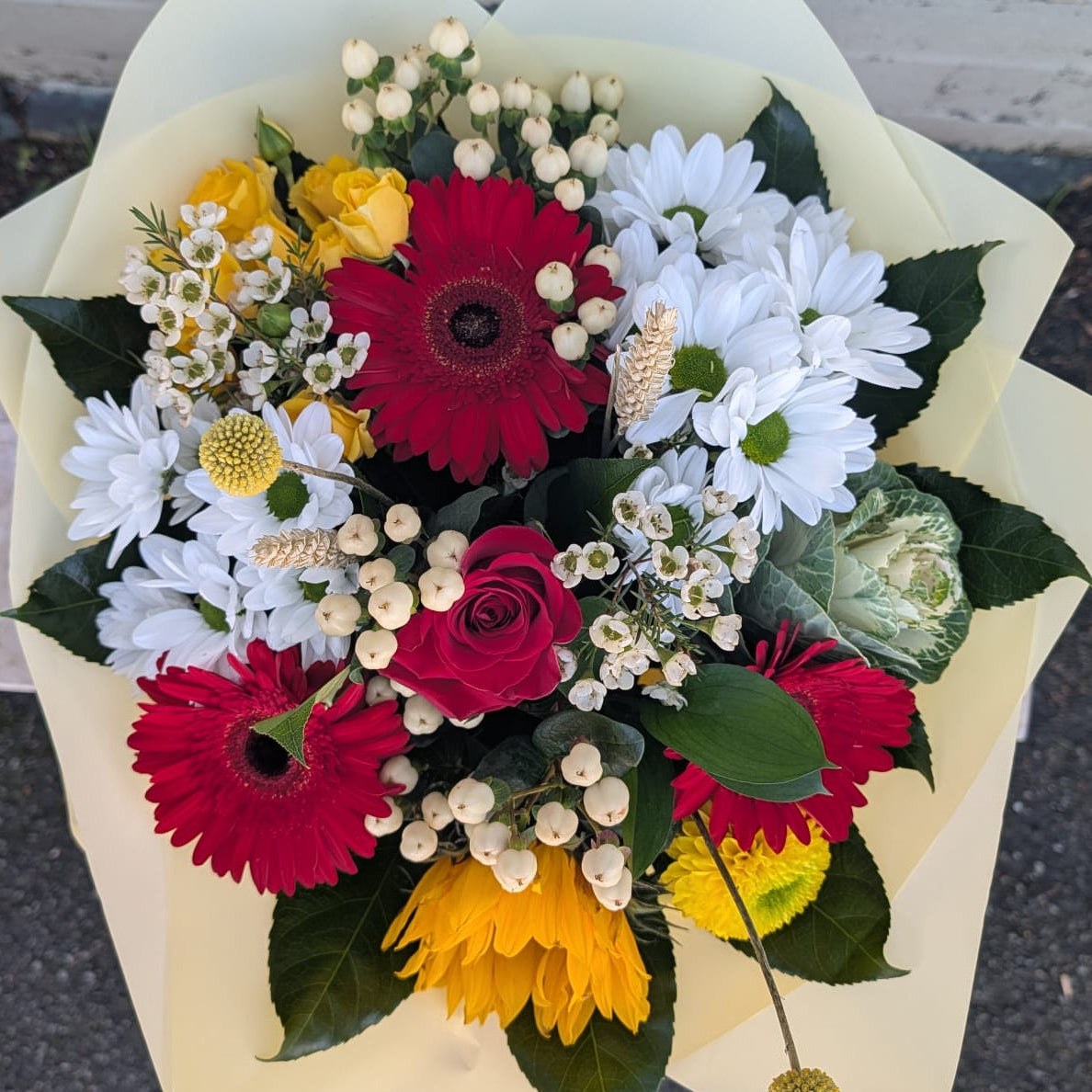 A vibrant mixed flower bouquet wrapped in pale yellow paper, featuring red and yellow gerbera daisies, white daisies, red roses, baby's breath, and green leaves, photographed outdoors against a white brick wall and asphalt ground.