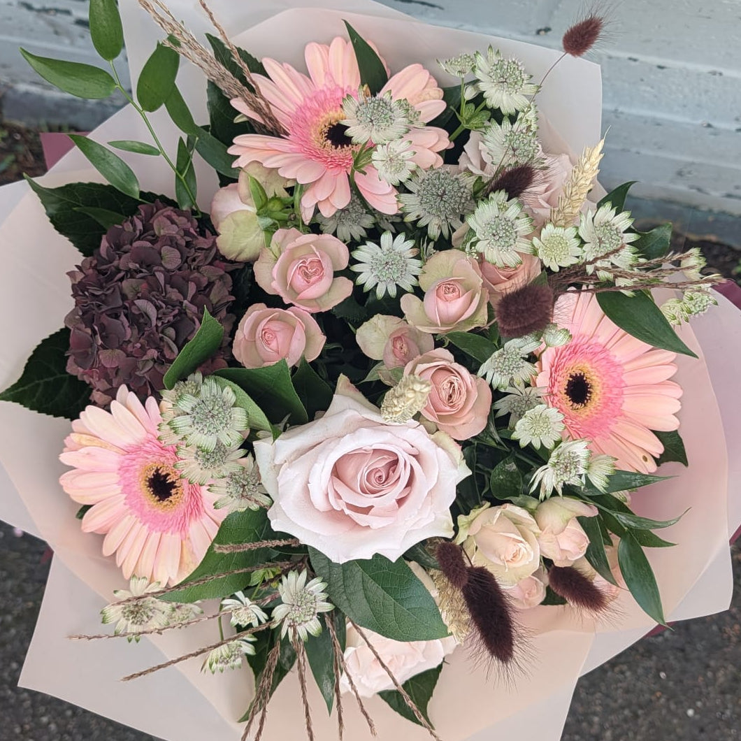 A hand-picked floral bouquet featuring pink roses, pink and white gerbera daisies, a dark purple hydrangea, and green foliage, wrapped in white paper and placed on asphalt next to a white brick wall.