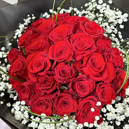 A large bouquet of vibrant red roses arranged in a circular pattern, surrounded by delicate white baby's breath and green stems, wrapped in dark gray paper.