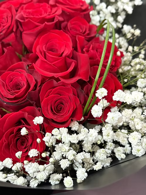 A close-up of a flower bouquet featuring vibrant red roses and delicate white baby's breath, arranged in a dark container.