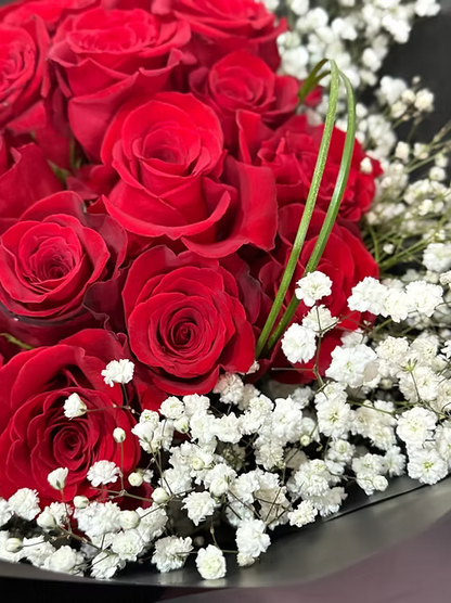 A close-up of a flower bouquet featuring vibrant red roses and delicate white baby's breath, arranged in a dark container.