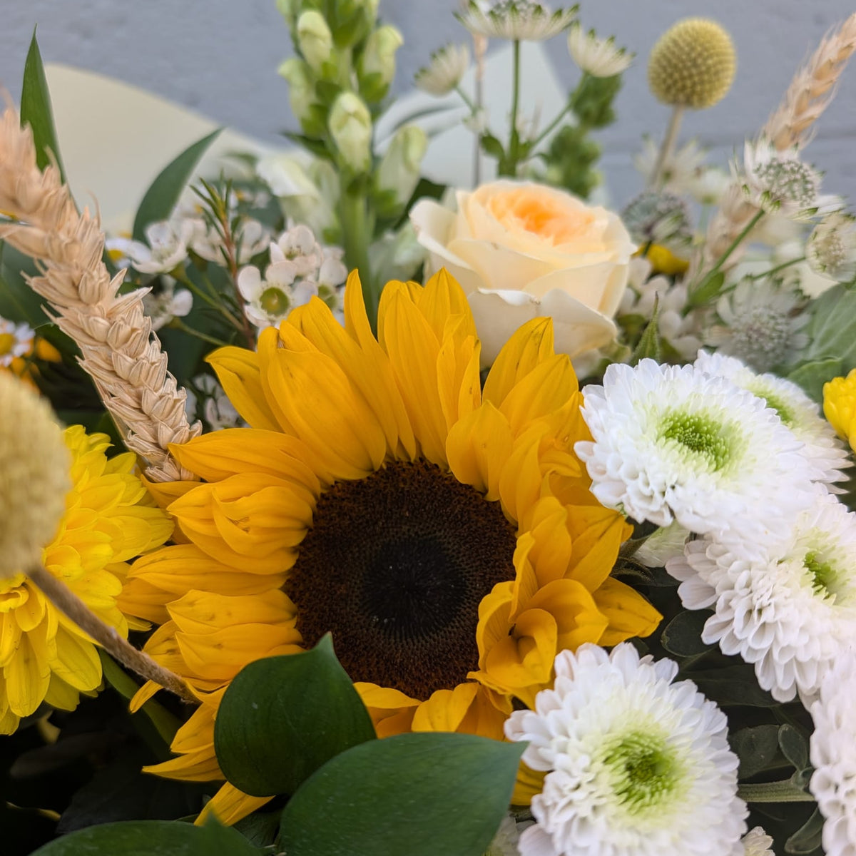 A close-up of a floral arrangement featuring a large yellow sunflower, white daisy-like flowers, green foliage, and a pale peach rose, photographed against a light gray background.
